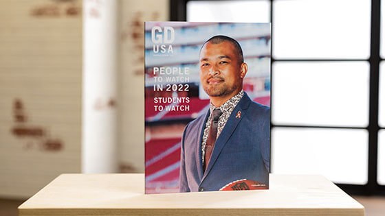 A Graphic Design USA magazine stands upright on a desk in a backlit room. On the cover, a man wearing a blue suit with a pink ribbon pin and holding an NFL football smiles for the camera. The cover reads, People to Watch in 2022, Students to Watch.