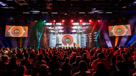 A person wearing a suit speaks on a stage in front of a large crowd. There is a large screen with a logo that says Hall of Fame XII behind them. Red, blue, and white lights illuminate the room.