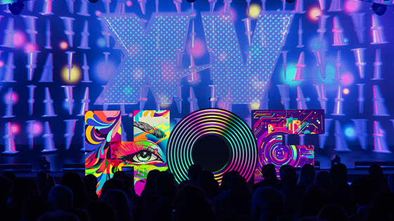 A large sculpture of the letters “HOF” sits atop the Full Sail Live Venue stage with different multicolored images projected onto each letter. The letters are in front of a large purple screen featuring the Roman numeral “XV” in large font. In front of the stage is a black silhouette of the crowd.