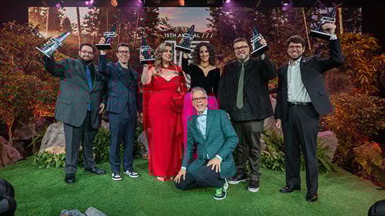 The Hall of Fame 15 inductees raise their awards in the air on a stage decorated with foliage. Garry Jones kneels in front.