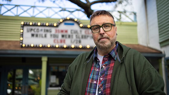 Aaron B. Koontz stands in front of a movie theater. He is wearing green glasses, a green sweatshirt, and a collared plaid shirt.