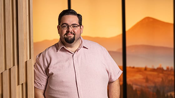 A man with glasses and a beard in a pink button-down stands next to a wooden panel, with a warm, mountainous sunset background behind him.