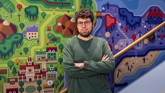 A bearded man in glasses and a green shirt stands with his arms crossed in front of a colorful, game-inspired illustrated mural.