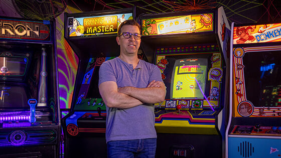 Jonathan Mayer smiles and stands in front of a row of arcade game machines for ‘Tron’, ‘Kung-Fu Master’, ‘Dig Dug’, and ‘Donkey Kong’.