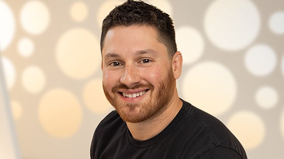 A headshot of a man with short brown hair and a black t-shirt smiling against a white background.