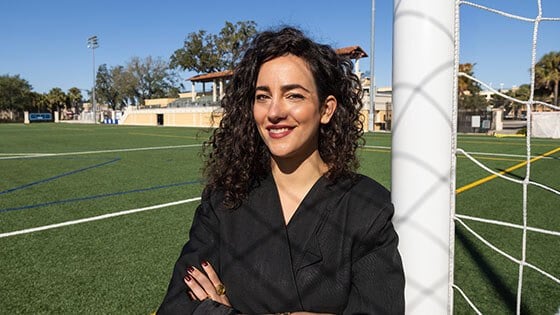 A woman with curly hair and a black jacket stands confidently by a soccer goalpost on a bright, sunny day at an outdoor field.