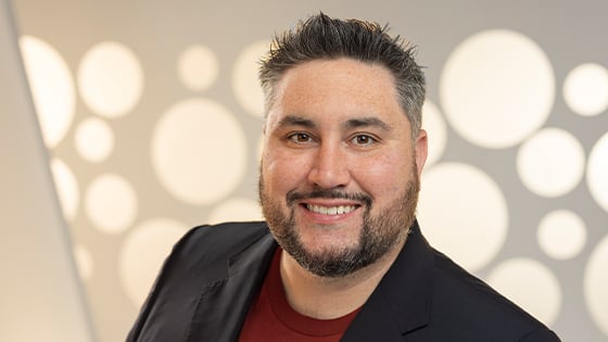 Michael La Plante smiles at the camera. He is wearing a red t-shirt with a black blazer and standing in front of a white backdrop.