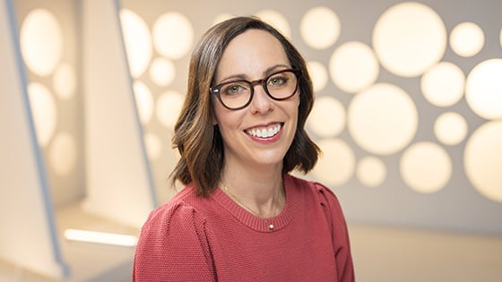 Nina Lopez-Corrado smiles at the camera. She is wearing brown glasses and a red sweater and standing in front of a white backdrop.