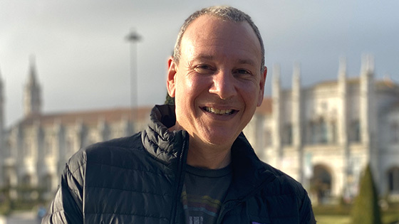 Sean Spuehler stands outdoors in front of a large building. He is smiling and wearing a black jacket and a blue t-shirt.