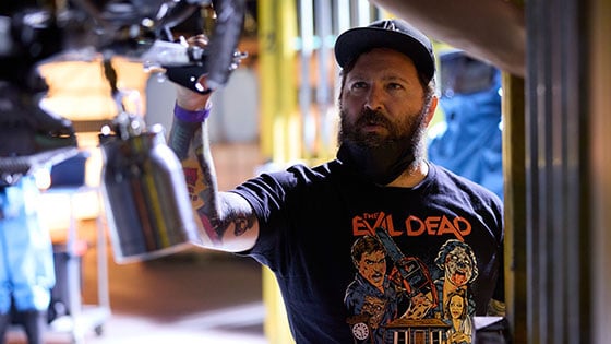 Steven C. Miller adjusts a piece of machinery on a science lab set. He is wearing an Evil Dead t-shirt.