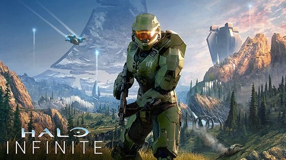 Halo main character Master Chief in signature green armor featuring the number 117 on his left chest plate in white, centered in front of a large grey triangular space ship with the words ‘Halo Infinite’ stylized in the bottom left corner.