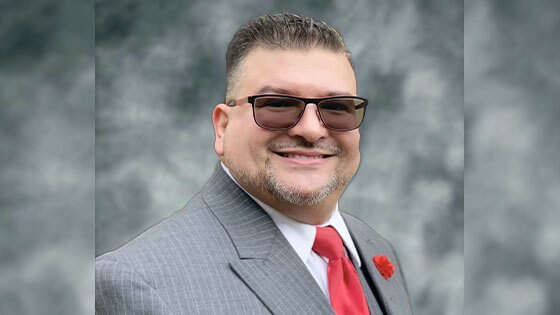 Will Torres wears a gray pinstriped suit with a red tie, a red carnation on the lapel, and sunglasses.