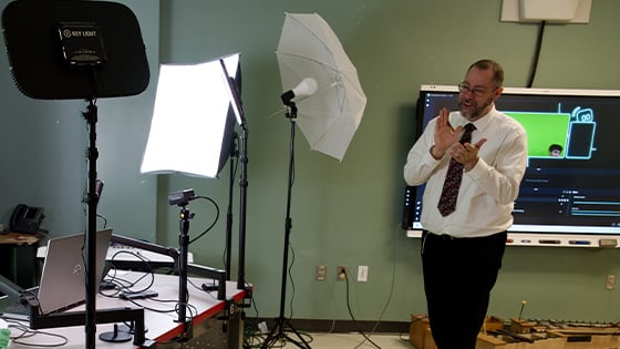 An instructor gestures while teaching in a studio setup with lights, cameras, and a green screen projection in the background.
