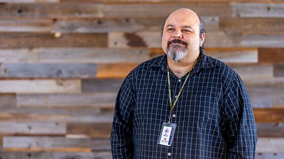 Instructor Eric Berzins, a man with a grey goatee, standing in front of a wood paneled wall wearing a blue button down dress shirt and green Full Sail lanyard with name badge.