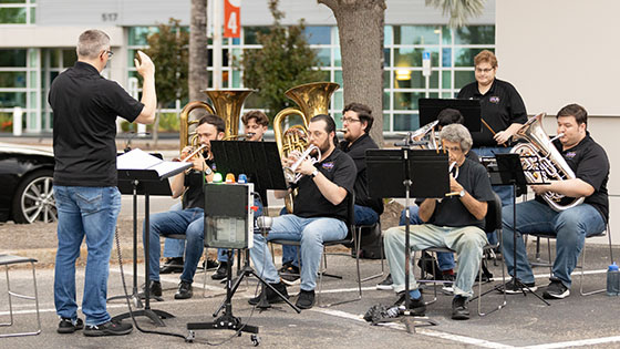 Members of the Brass Band of Central Florida perform in a parking lot, sitting down and reading from their music stands while a conductor stands at the front.