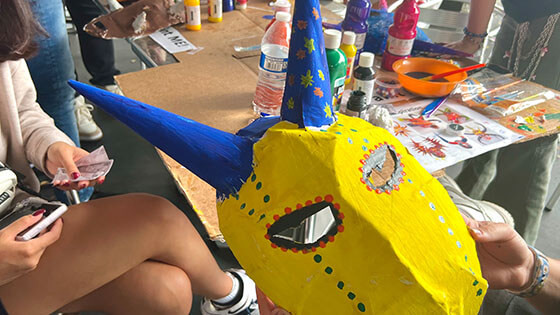 A person holds up a hand painted horned mask in front of a crowded arts and crafts table.