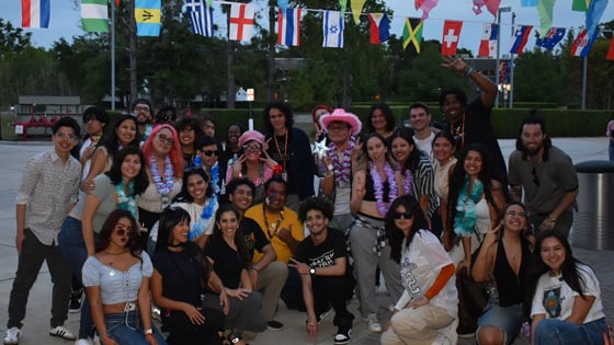 A diverse group of young adults poses outdoors under international flags, dressed festively with leis and fun accessories.