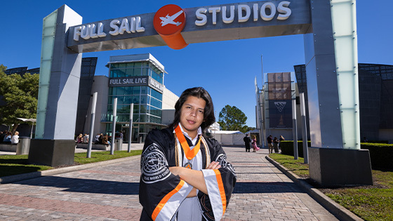 John Tobar stands with his arms crossed in front of the Full Sail Studios archway. He is wearing a graduation gown and an Armada medallion.
