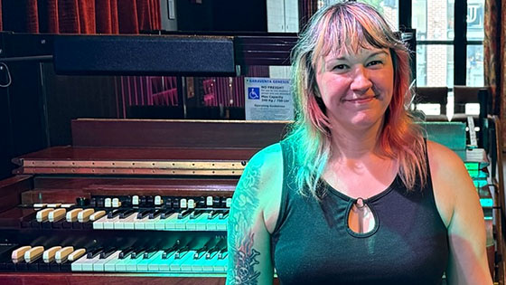 Kim Kozak sits on a piano bench in a backstage area at Garcia’s. She is smiling and wearing a black dress and tights.