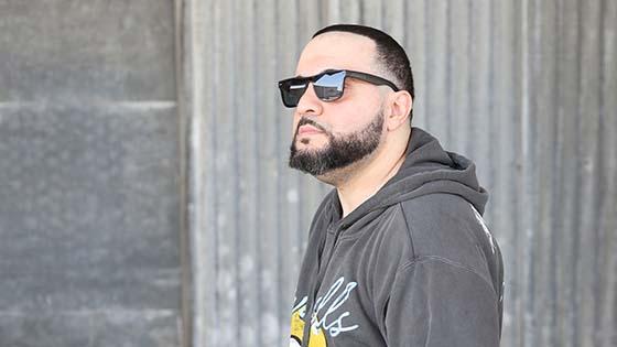 Full Sail grad George Doman wearing black sunglasses and a grey pullover hoodie with a yellow and light blue design, standing in front of a large stone wall while looking off into the distance.