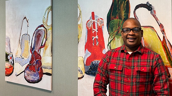 Anthony Roberts Jr. stands in front of two paintings of shoes. He is wearing black-rimmed glasses and a plaid button-down shirt.