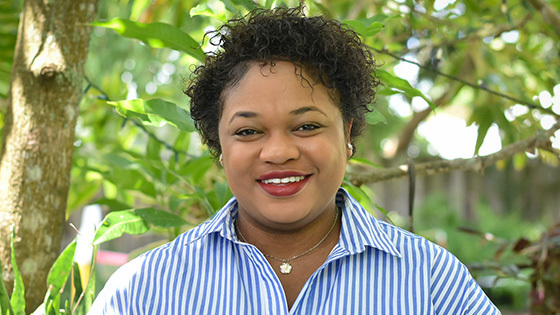 Jessica stands in front of a lush garden, surrounded by greenery. She is wearing a blue pinstripe shirt and is smiling.