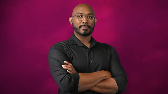 Gregg is wearing a black shirt and rectangular glasses. He is standing in front of a pink background with his arms crossed.