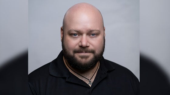 Grad Corey Guerra is wearing a black polo shirt and posing for a headshot against a light grey backdrop.