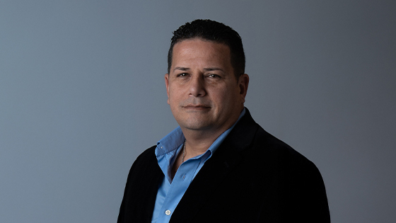 Sandy Navarro smiles at the camera in front of a grey background. He is wearing a blue button-down shirt and a black blazer.