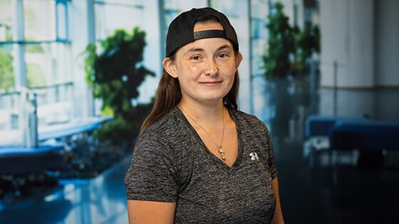 A person wearing a backward cap and a gray Under Armour shirt smiles in a bright, modern indoor space with plants in the background.