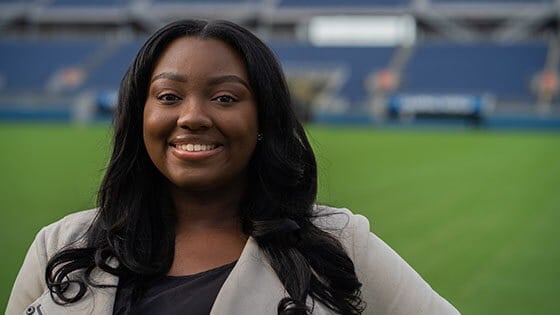 A woman with long brown hair smiling in a beige blazer while standing on a soccer pitch.