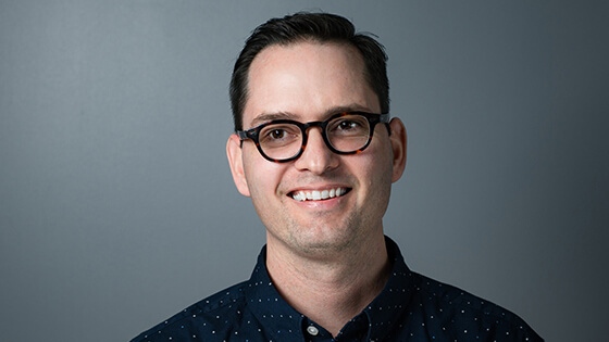 Digital Arts & Design grad Paul Shank wearing a blue, polka-dotted shirt in front of a gray background.