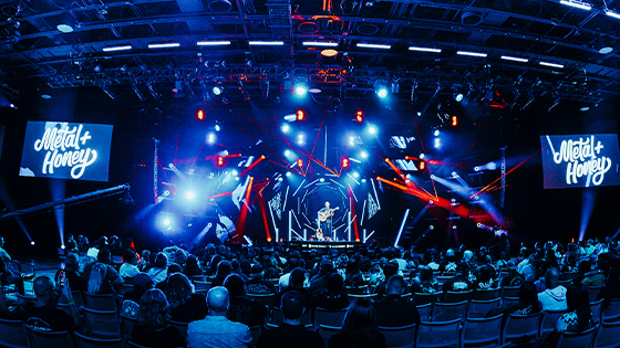 A vibrant charity concert featuring Matt Heafy of Trivium on stage with dynamic lights, a packed audience, and 'Metal + Honey' branding on screen.