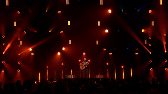 Trivium lead-singer Matt Heafy on a dimly-lit stage playing an acoustic guitar in front of a large crowd.