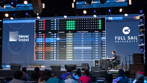 A person is seated at a desk on a stage in front of a crowd of students while a large LED screen behind displays a mirror of the audio program running on their computer.