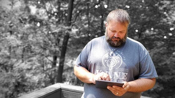 Grad Ryan Backa in a blue shirt with a white dragon design, holding an electronic tablet in front of a wooded area in greyscale.