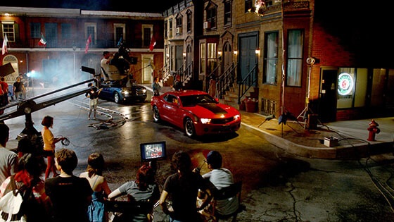 Full Sail University's Backlot in the evening filled with a full film production crew shooting a scene featuring two muscle cars, one red and one black.