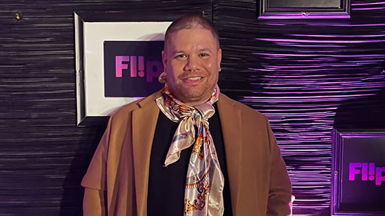 Javi Simons stands in front of a purple wall. He wears a black t-shirt, jeans, a brown overcoat, and a colorful scarf. He is smiling.