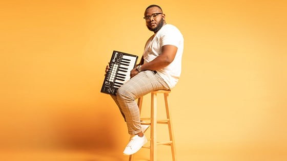 Chavez Parker sits on a stool and holds a keyboard. He wears glasses, a t-shirt, brown pants, and sneakers.