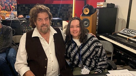 Alan Parsons sits with Steffie Tjandra in front of a mixing board in a recording studio. They are smiling at the camera.