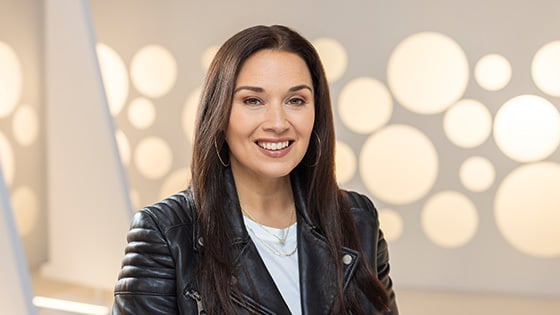 A headshot of a woman with straight brown hair and hoop earrings smiling while wearing a leather motorcycle jacket against a white background.