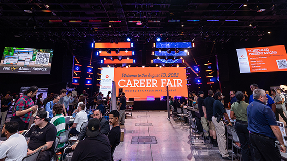 A bustling career fair with several booths set up. In the center, a giant LED screen welcomes guests to the Career Fair.