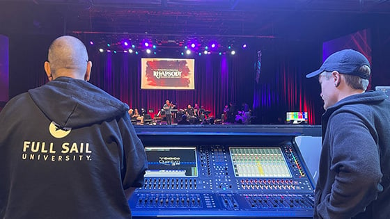 Two people's backs are facing the camera as they overlook an audio console and stage at The Plaza Live.