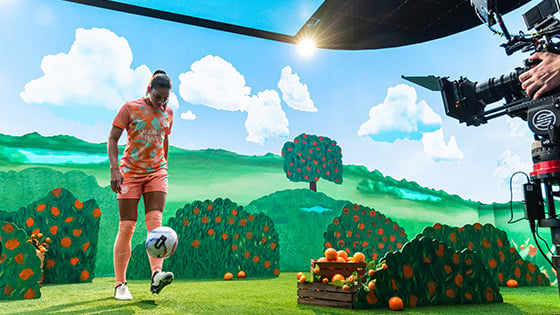 An Orlando Pride player kicks a soccer ball toward a camera on a film set that features an animated orange grove.