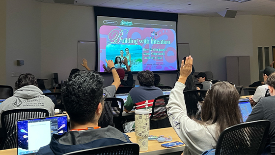 Students sit in an audience facing speakers Sofia Adamary and Gabriella Pedraza. A few students have their hands raised. A projector screen displays “Building With Intention” and a picture of the two speakers.