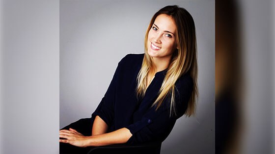 Grad Lisa Maskey, a smiling woman with straight blond hair wearing a black three-quarter sleeve top seated against a grey backdrop.