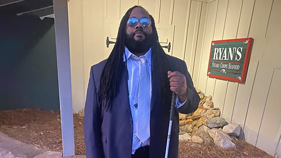 Andre Woods stands outdoors in front of a restaurant. He is wearing a blue suit and sunglasses. He is holding a mobility cane.