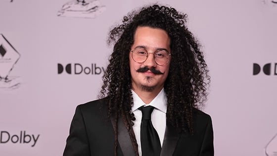 Guillermo Moya smiles at the camera in front of a white background. He is wearing a black suit, black tie, and glasses.