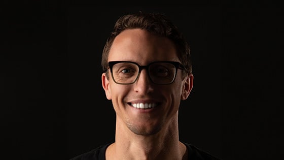 Drew Becker smiles at the camera in front of a dark background. He is wearing black glasses and a black shirt.