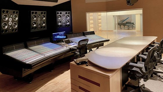 A conference table faces a wall with mixing boards and speakers; a recording area with a grand piano is in the background.